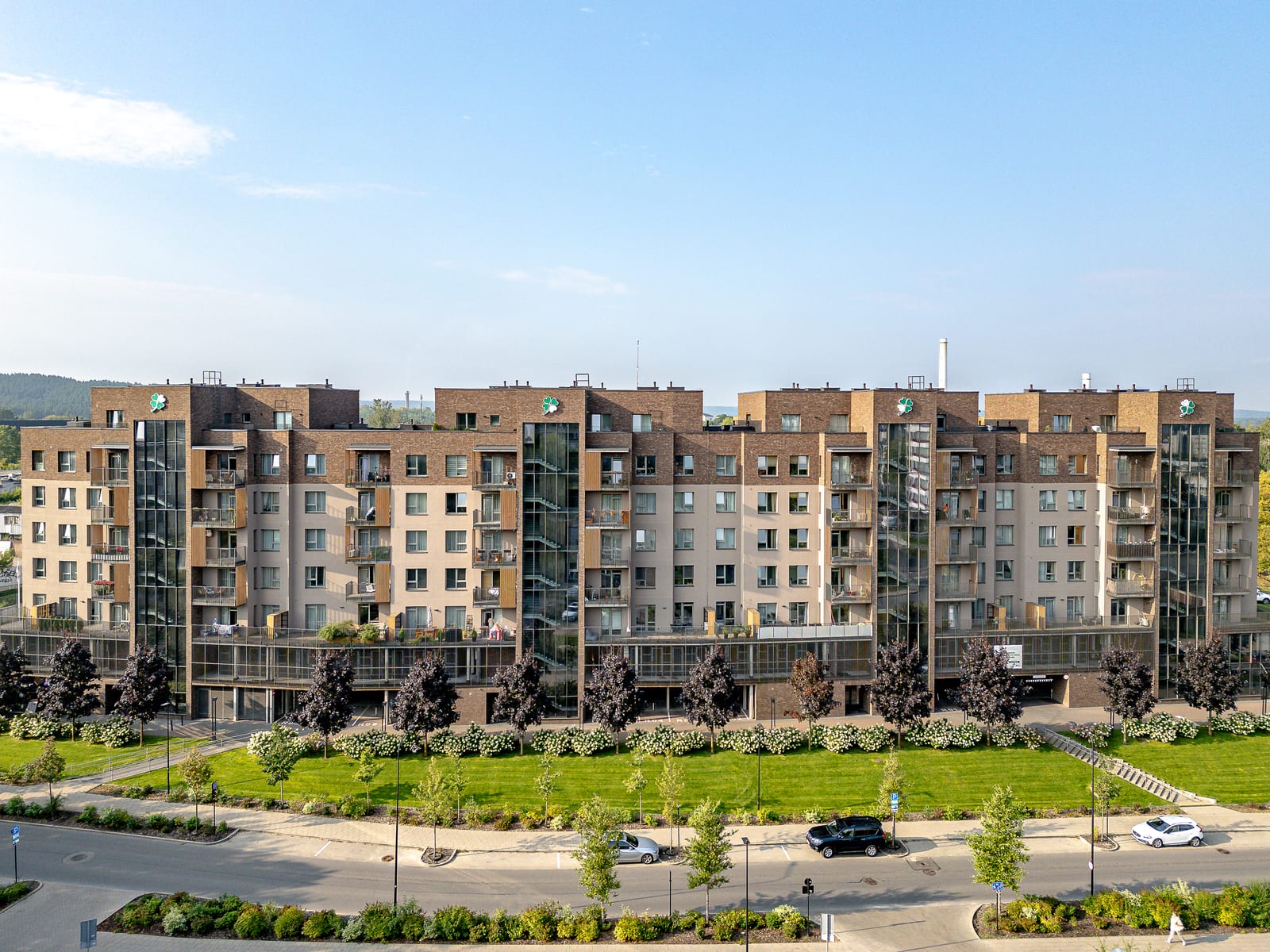 Lucky Homes by SAVO in Vilnius from the air: the whole clinker-brick and plaster building with its glazed stair towers, a band of white hydrangeas, lawn and the street in front.
