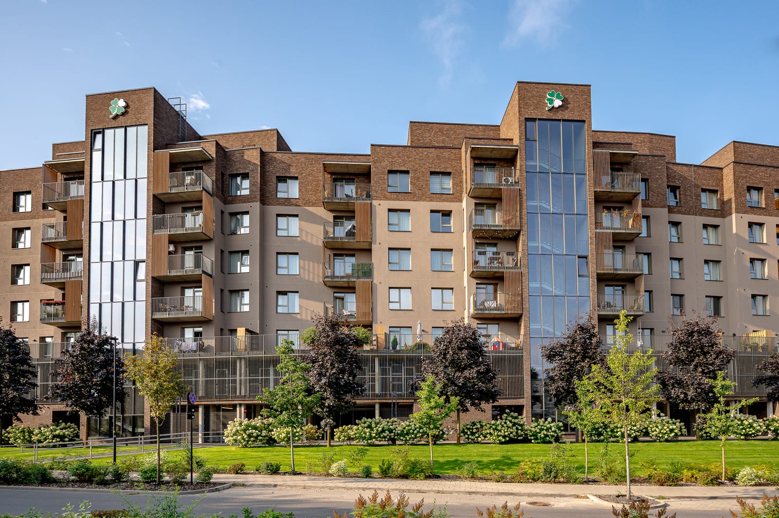 The facade from the drive: two glazed stairwells with the clover emblem, balconies with timber slats, hydrangeas and lawn.