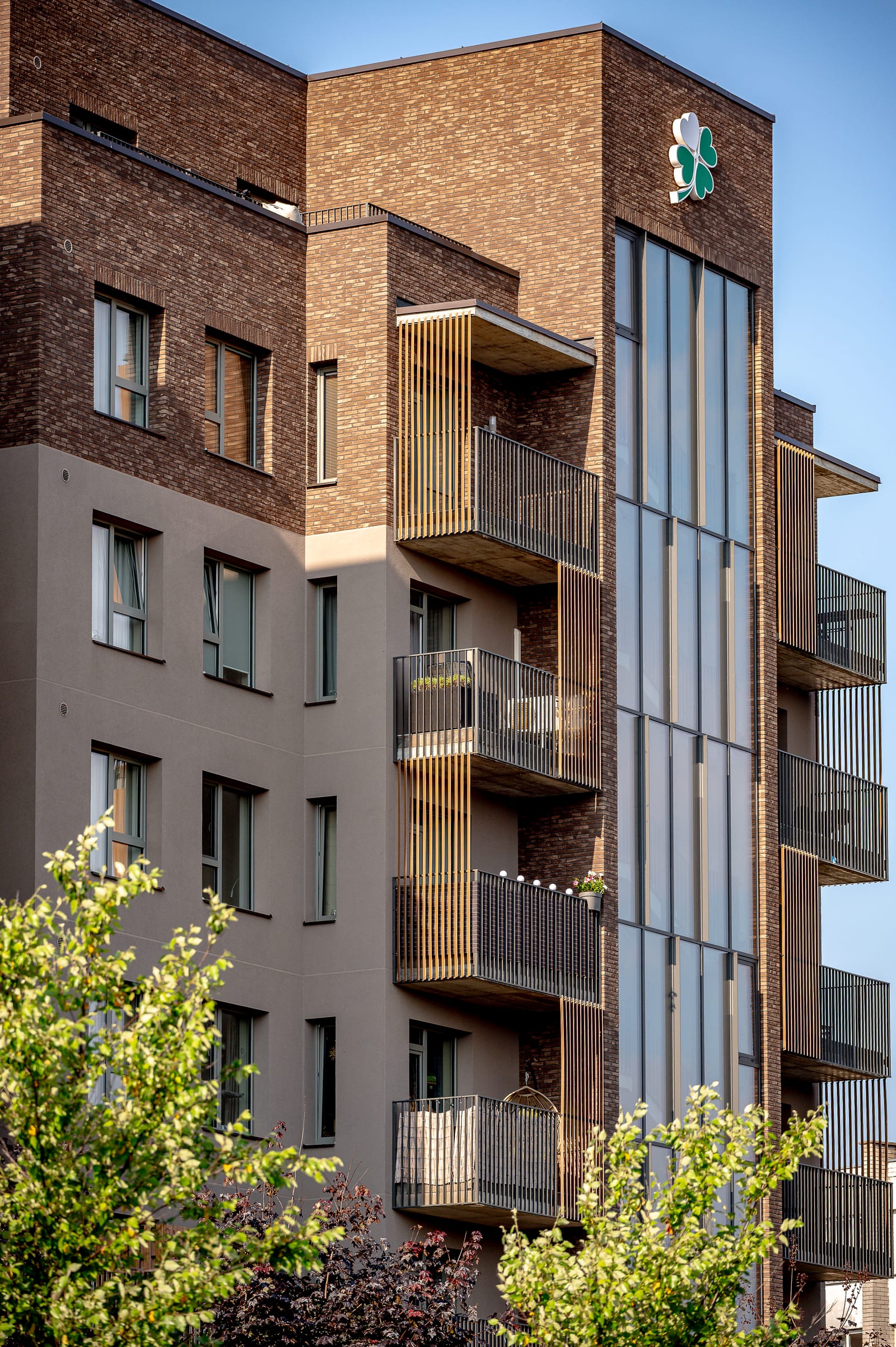 The corner of the building from below: the glazed stairwell with the clover emblem, clinker brick, a plastered wall and balconies with timber slats.