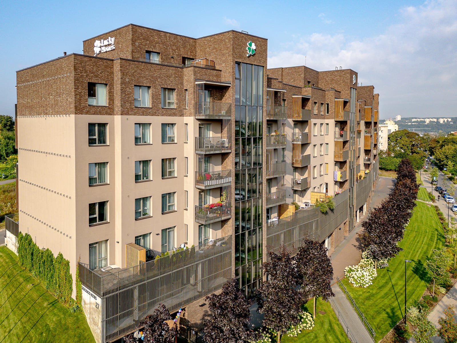 The corner of the building from a drone: the three-dimensional Lucky Homes lettering and the clover emblem on the parapet, with the concrete wall carrying the street name, a hedge and an avenue of maples below.