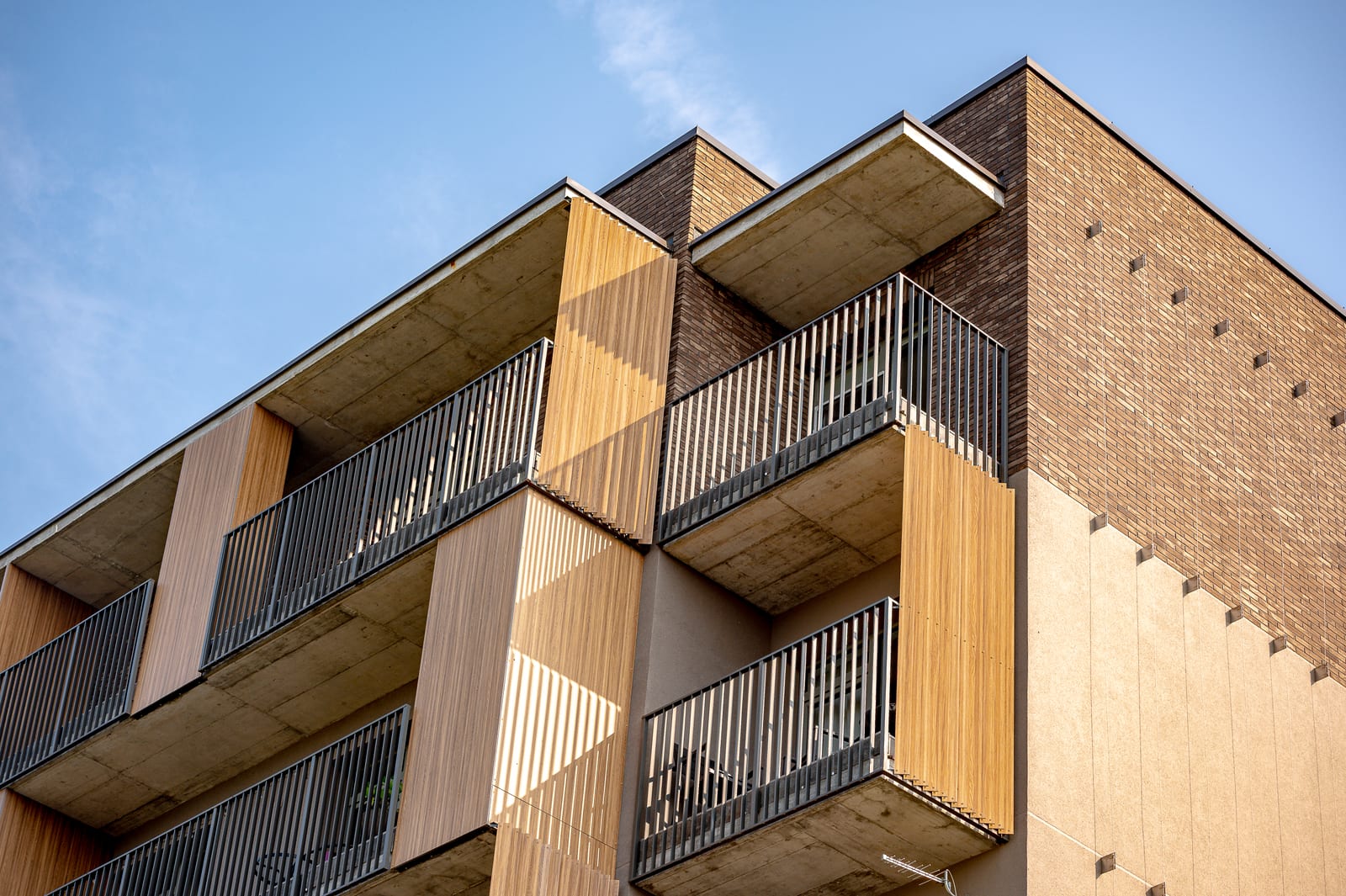 An architectural detail: the corner of the building from below — concrete balcony slabs, timber slats and clinker brickwork against the sky.